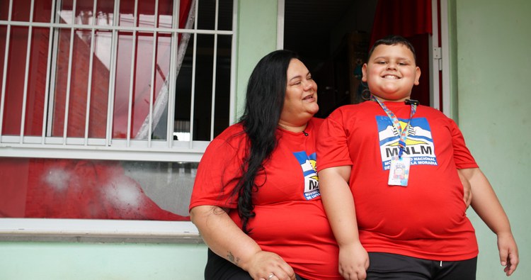 Na imagem mulher ao lado de seu filho com autismo comemorando sua casa própria